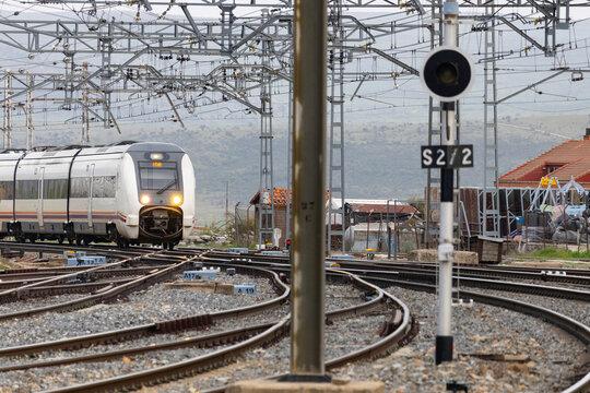 Commuters and travellers gather along the platforms at &Aacute;vila railway station as a regional train arrives, framed by overhead cables and urban architecture