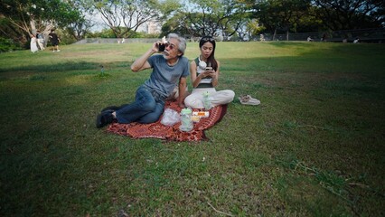 Fototapeta premium Senior man making phone call and young woman using social media during picnic in park