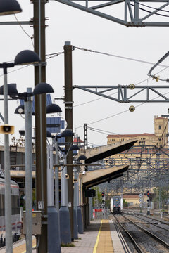 Commuters and travellers gather along the platforms at &Aacute;vila railway station as a regional train arrives, framed by overhead cables and urban architecture