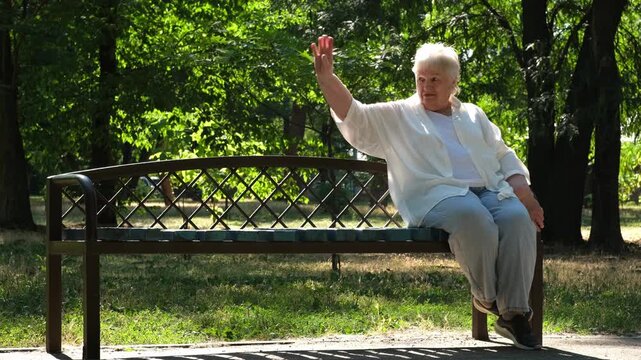 Happy smiling senior woman sitting on bench in city park on sunny summer day. Retired elderly lady resting outdoors and waving to someone. Retirement concept