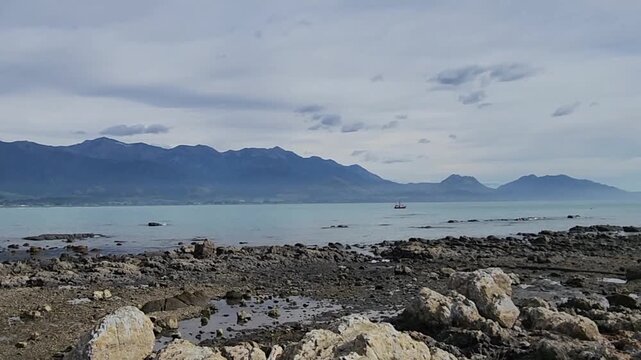 Kaikōura, New Zealand &mdash; Rocky Coastal Shoreline with Calm Blue Water, Low‑Tide Pools, Scattered Stones and a Distant Mountain Range Beneath a Soft, Partly Cloudy Sky in a Peaceful Natural Landscape