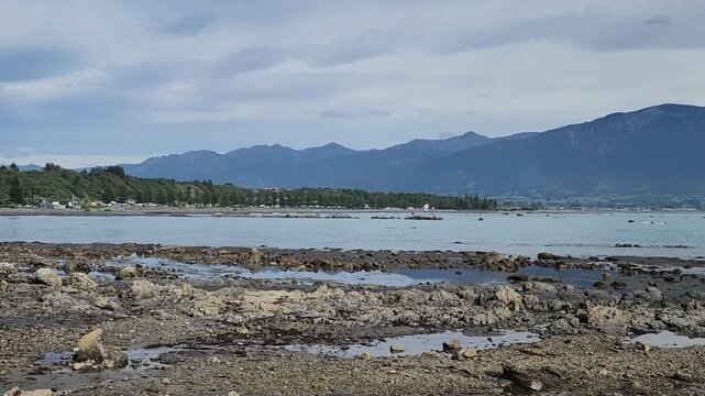 Kaikōura, New Zealand &mdash; Rocky Coastal Shoreline with Calm Blue Water, Low‑Tide Pools, Scattered Stones and Forested Mountain Range Beneath a Soft, Partly Cloudy Sky in a Peaceful Natural Landscape
