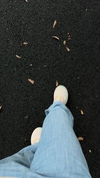 Handheld tracking shot of feet walking forward on asphalt path with scattered leaves in park during daytime autumn casual stroll