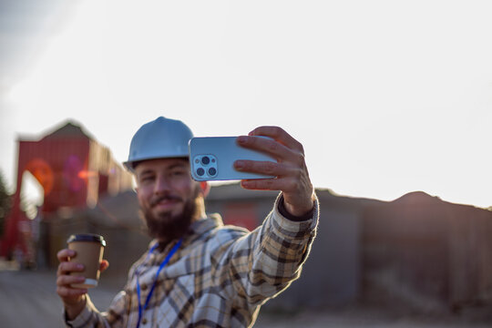 Construction worker making selfie with smartphone and coffee, documenting work progress at industrial site