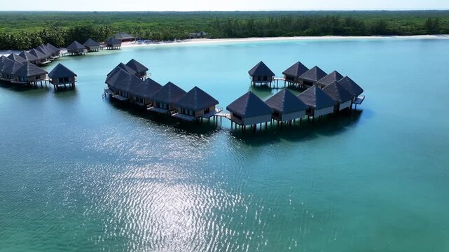 Aerial tracking shot of over water bungalows and villas gliding above turquoise lagoon along tropical coastline beach at midday