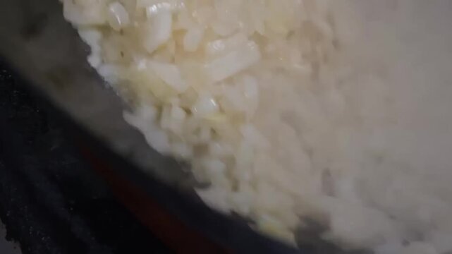 High-angle, close-up shot of finely diced white onions being saut&eacute;ed in a metal pot. The onions appear translucent and soft, typical of the initial "soffritto" or base stage of a recipe.