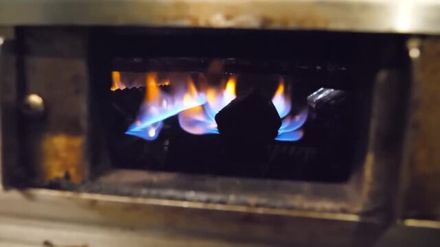 A high-definition, close-up stock video of a powerful blue and yellow flame from a torch or burner igniting wood or charcoal inside an industrial metal container. The intense heat and fire create a dy
