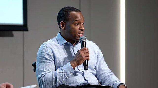 This features a confident African American man delivering a presentation in a modern conference room. He speaks into a microphone with authority and conviction, engaging his audience with a compelling