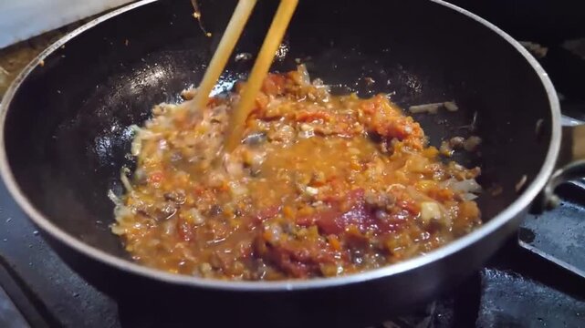 Close-up footage of red curry paste being saut&eacute;ed with chopped onions and oil in a black frying pan on a gas stove. A chef uses wooden chopsticks to stir the ingredients as they sizzle and release fra