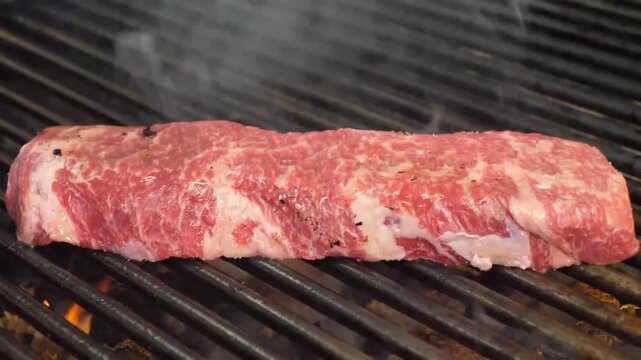 Close-up footage of a raw, well-marbled beef skirt steak being brushed with oil or marinade while sizzling on a hot, cast-iron grill over open flames. The steak is prized for its deep, beefy flavor an