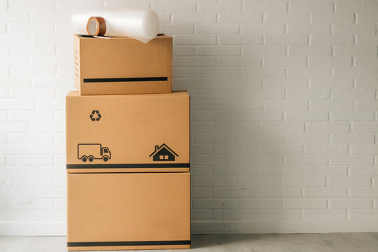 Stack of cardboard moving boxes with truck and house icons, bubble wrap, and packing tape on top, against a white brick wall. Perfect for relocation and moving concepts