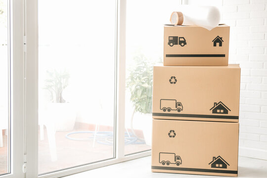 Stack of cardboard moving boxes with truck and house icons, bubble wrap, and packing tape on top, against a white brick wall. Perfect for relocation and moving concepts