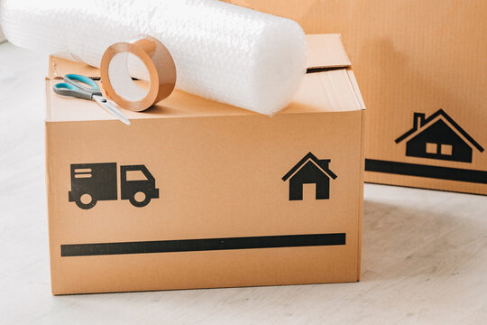 Stack of cardboard moving boxes with truck and house icons, bubble wrap, and packing tape on top, against a white brick wall. Perfect for relocation and moving concepts