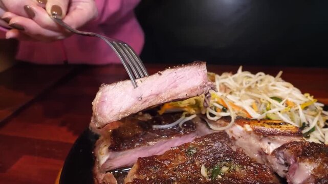 close-up shot of a person using a fork to cut into a perfectly cooked, medium-rare T-bone steak served on a black sizzling plate with a side of fresh salad. The rich, savory meat juices and vibrant si