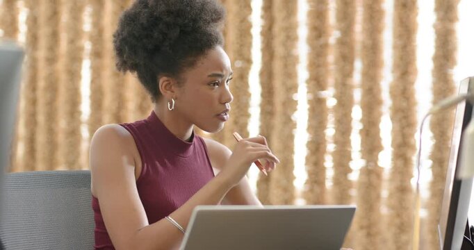 African American woman in burgundy typing on laptop, checking monitor for work, holding pen at desk