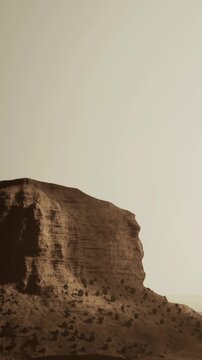 A group of rocky formations stands tall in a barren landscape. The sky is light and featureless, while the ground is covered with small stones and dust. The scene is vast and empty.