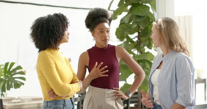 Diverse women at work shifting into smiles with left colleague touching sternum for camera by plant