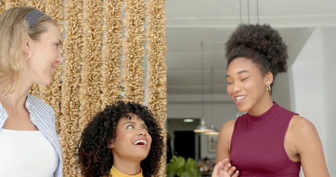 Diverse female friends in kitchen laughing pointing after seated woman raising hand near gold beads