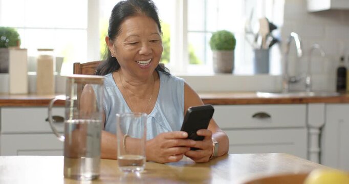 Senior Asian woman in blue blouse laughing and tapping phone at kitchen table after getting message