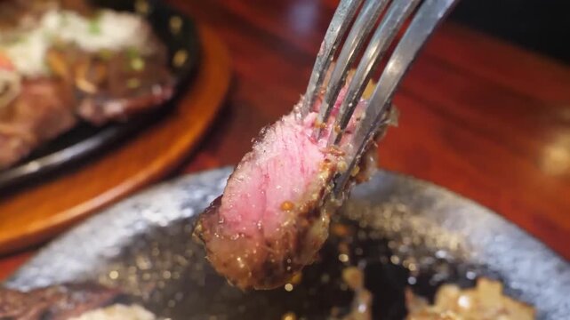 A close-up shot of a fork lifting a perfectly cooked, juicy, medium-rare steak slice from a sizzling hot plate. The rich red interior and seared exterior of the beef are clearly visible, highlighting 