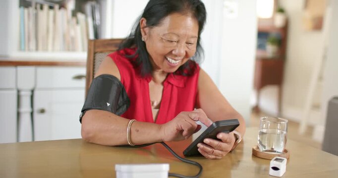 Senior Asian woman wearing red blouse touching base, tapping monitor with cuff at table checking BP