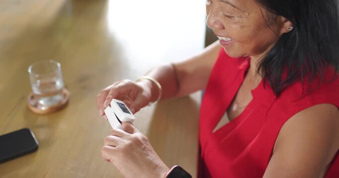 Senior Asian woman prompted by device picking up fingertip monitor at wooden table, reading results