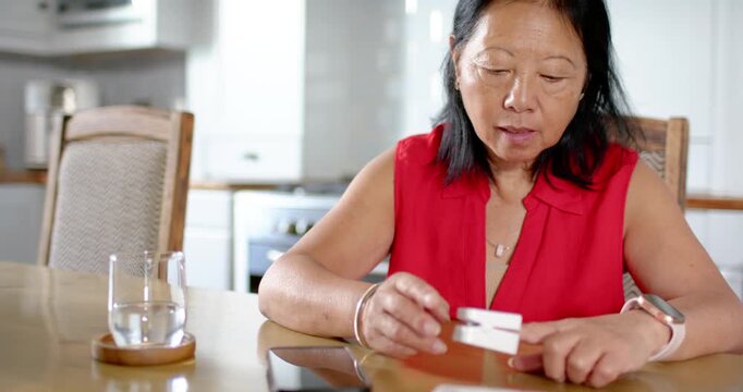 Senior Asian woman in red blouse checking pill holder, drinking water after reading card at kitchen