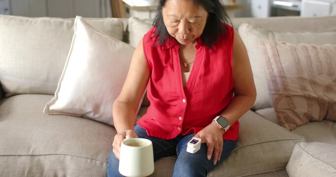 Senior Asian woman finishing sip, placing mug, clipping oximeter and checking oxygen level at home