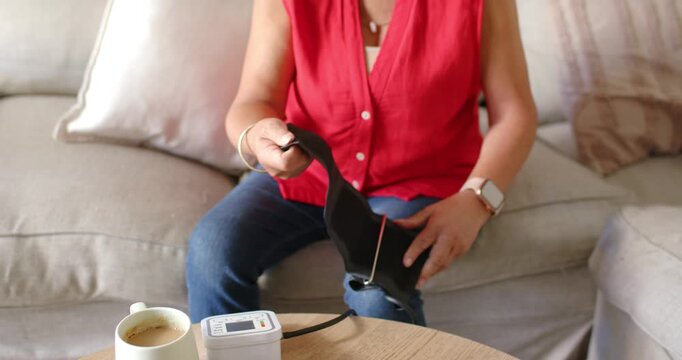 Senior woman on sofa picking cuff from table near monitor, unfolding, securing on arm for reading