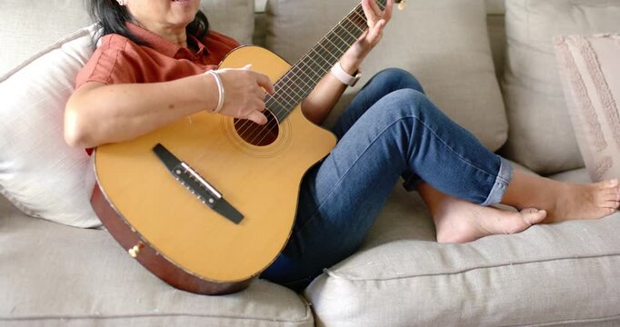 Senior Asian woman settling into sofa, strumming guitar and changing chords while practicing