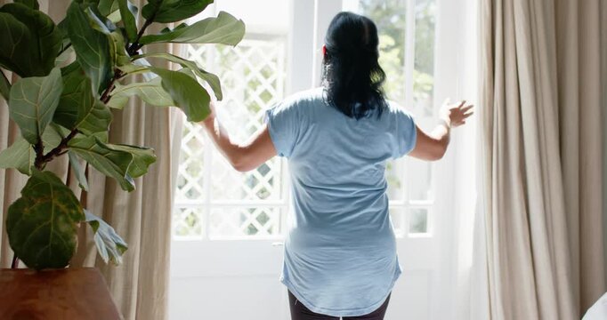 Senior woman reaching for drapes, pulling aside, stepping at door, showing sunlit lattice and plant