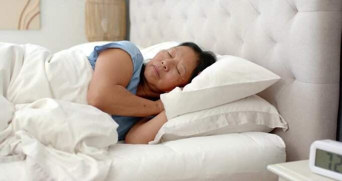 Senior Asian woman waking to alarm clock ringing, rubbing eyes and stretching arms overhead on bed