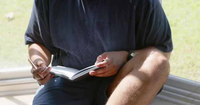 Pen poised on notebook, African American man in navy T-shirt journaling on window seat, glancing