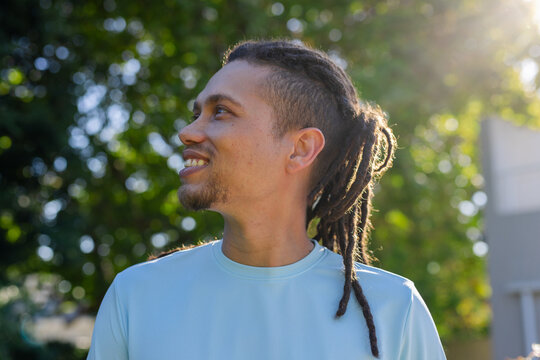 Adult man smiling while looking up outdoors, wearing light blue crew-neck T-shirt and dreadlocks