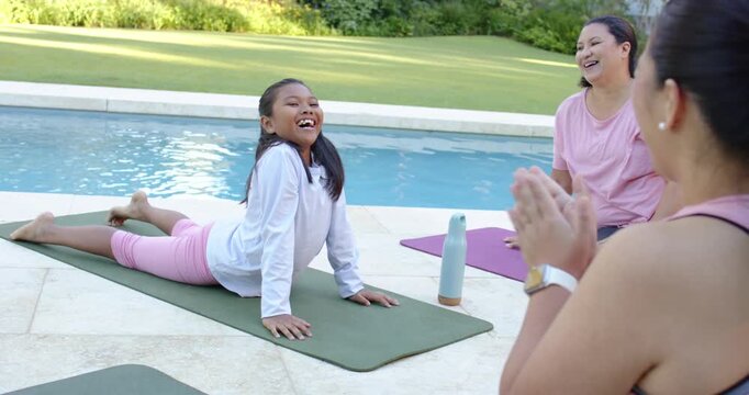 Diverse female friends clapping and encouraging youth holding cobra backbend on green mat by pool
