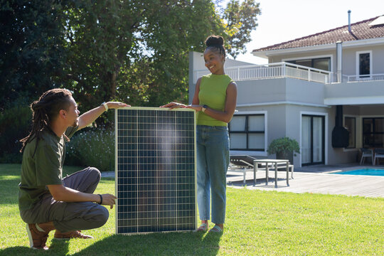 African American couple installing large photovoltaic solar panel on grassy lawn near pool