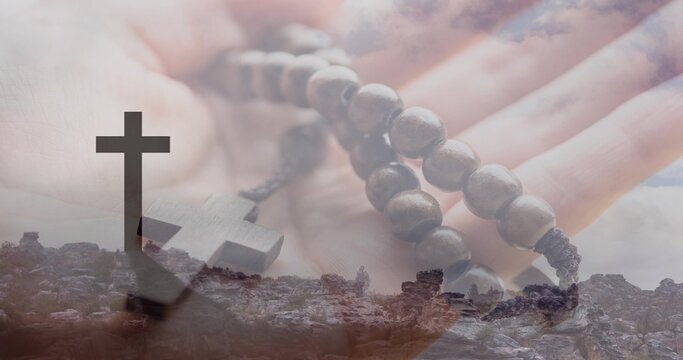 Holding open palm, adult hand cradling wooden rosary beads over rocky hilltop silhouetted cross