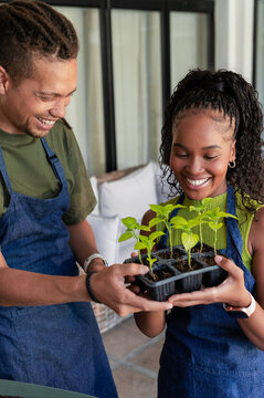 African American couple holding black plastic seedling tray on covered patio, wearing denim aprons