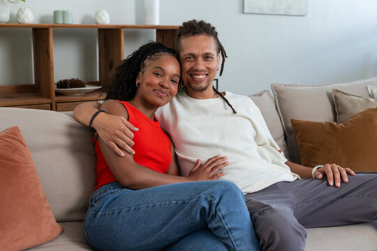 African American couple sitting closely on beige sofa in living room, showing red top and watch