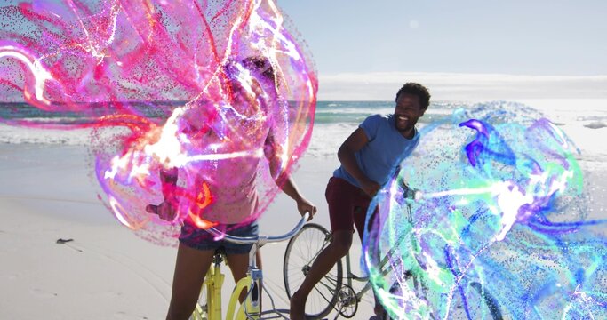 Laughing couple standing, straddling yellow, dark bikes on beach, in denim shorts, particle ribbons