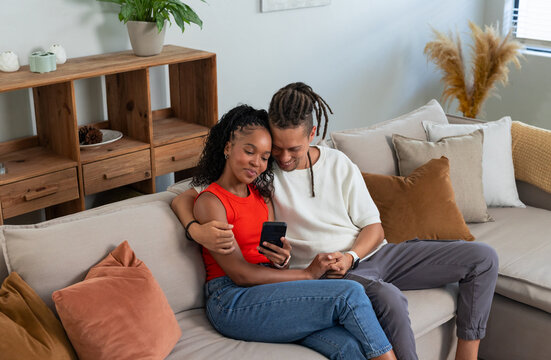 African American couple sitting on sectional sofa in living room sharing smartphone, rust pillows