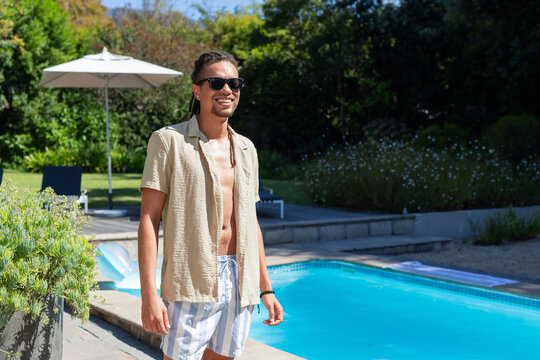 Man in sunglasses standing at pool edge wearing open beige shirt and striped trunks