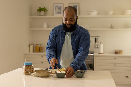 Bearded man wearing denim making breakfast at kitchen island, assembling granola bowl with bananas
