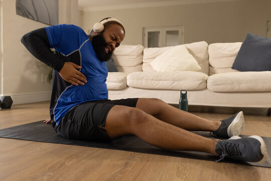 African adult man in workout gear with headphones clutching left side on mat in living room