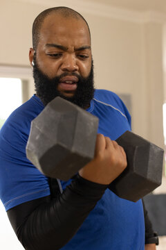 African American man curling hex dumbbell at home wearing blue shirt, sleeves, earbud, watch