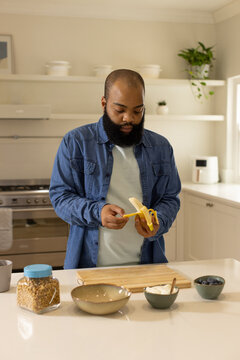 African American man wearing denim shirt peeling banana at kitchen island with cutting board