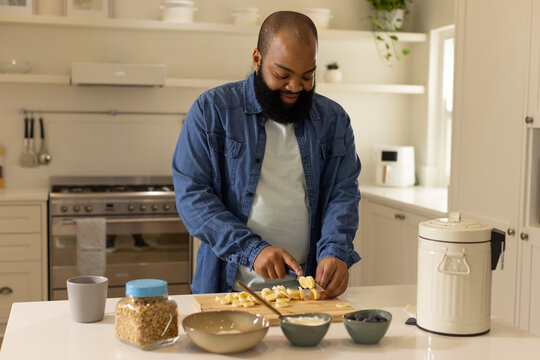 African American man slicing banana on wooden cutting board at kitchen island with granola jar