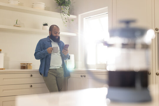 African American man in his 30s leaning against counter holding mug and checking phone in kitchen