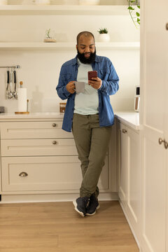 African American man leaning on kitchen counter, holding gray mug while checking red smartphone