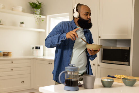 African man wearing denim shirt, headphones stirring bowl and spoon in kitchen with French press